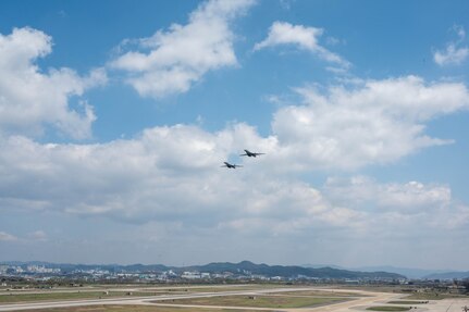 Two aircraft fly in close formation with a white, puffy clouds and a blue sky behind them, over a runway with a town in the distance.