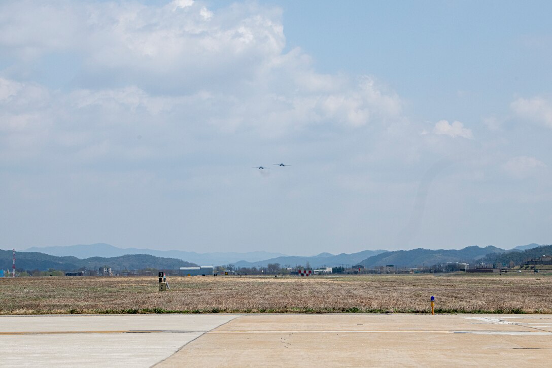 Two small aircraft appear in the gray-blue sky over a field and mountains in the background.