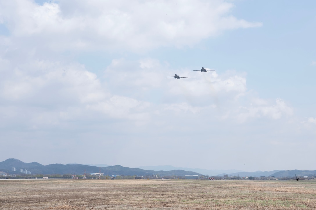 Two B-1 bombers fly in a gray blue sky over a grass field with mountains low in the background.