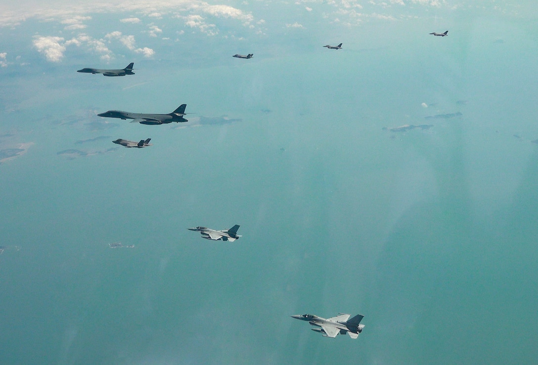 Five aircraft fly in a v-formation, viewed from above, over a blue-green sea with islands below.