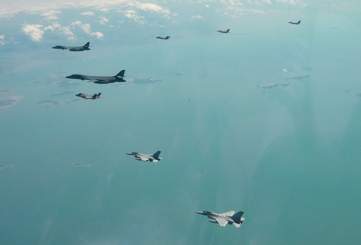 Five aircraft fly in a v-formation, viewed from above, over a blue-green sea with islands below.
