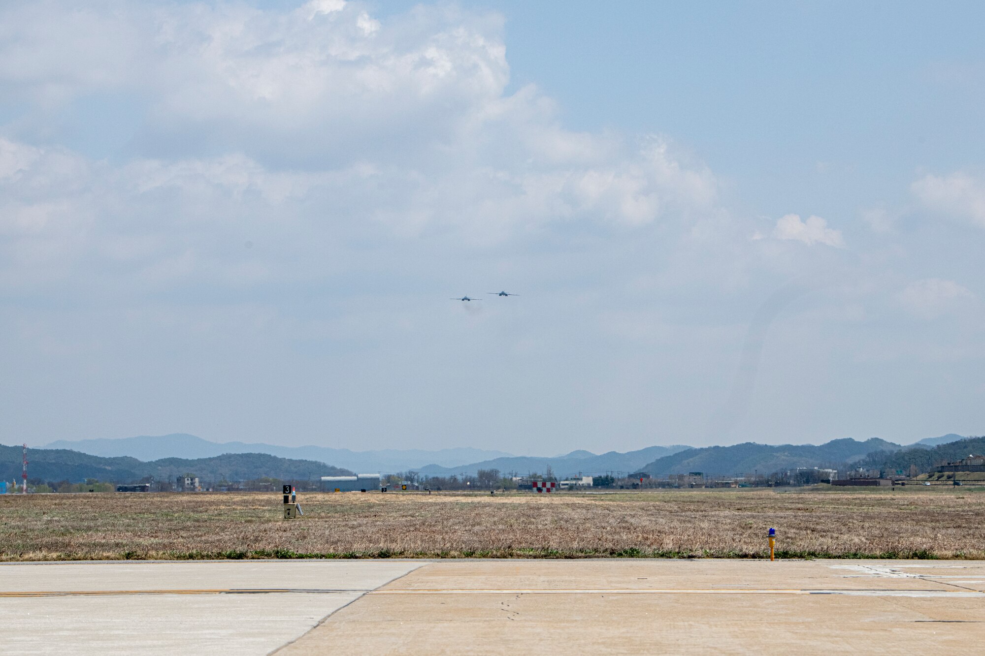 Two small aircraft appear in the gray-blue sky over a field and mountains in the background.