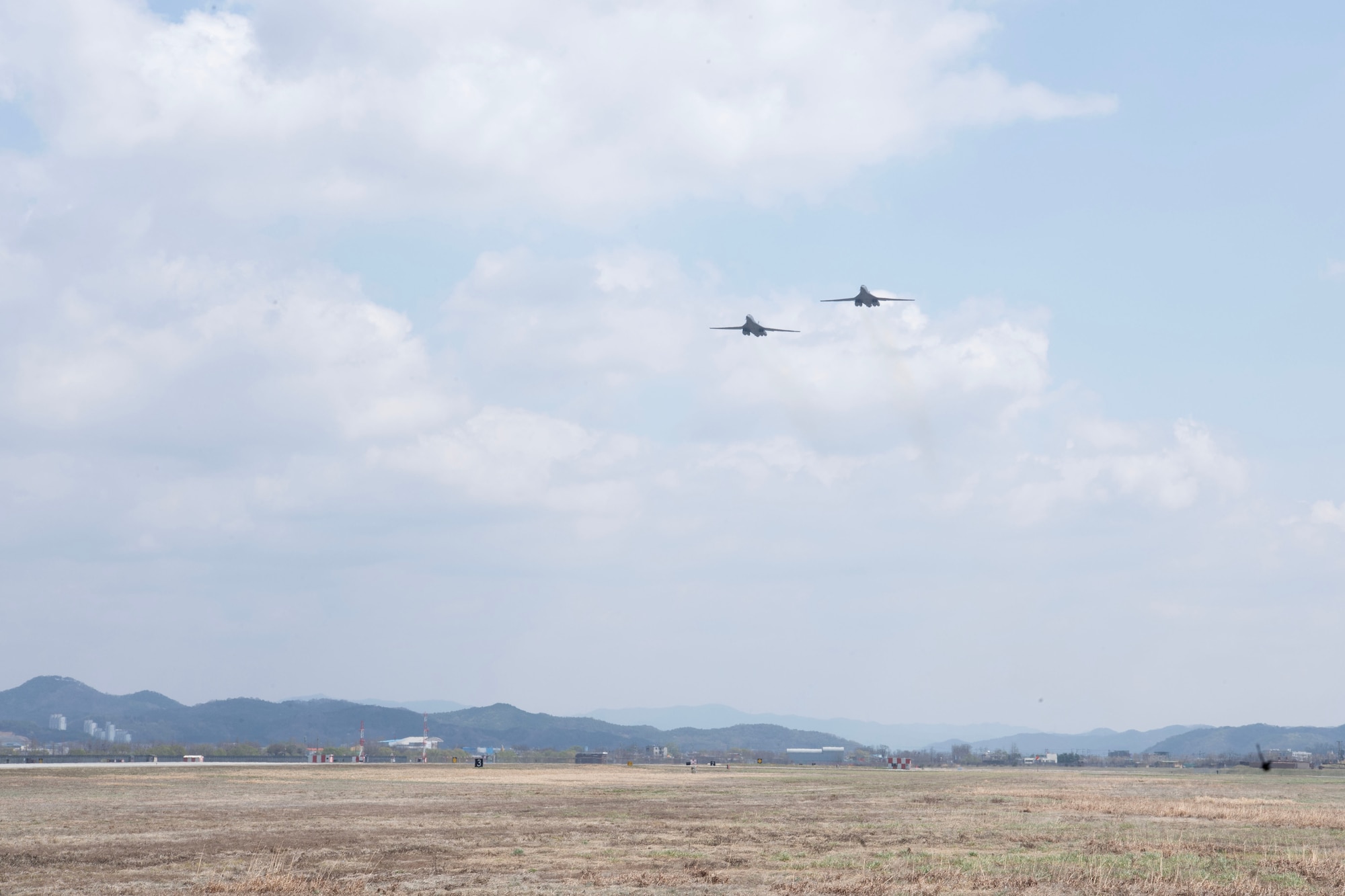 Two B-1 bombers fly in a gray blue sky over a grass field with mountains low in the background.