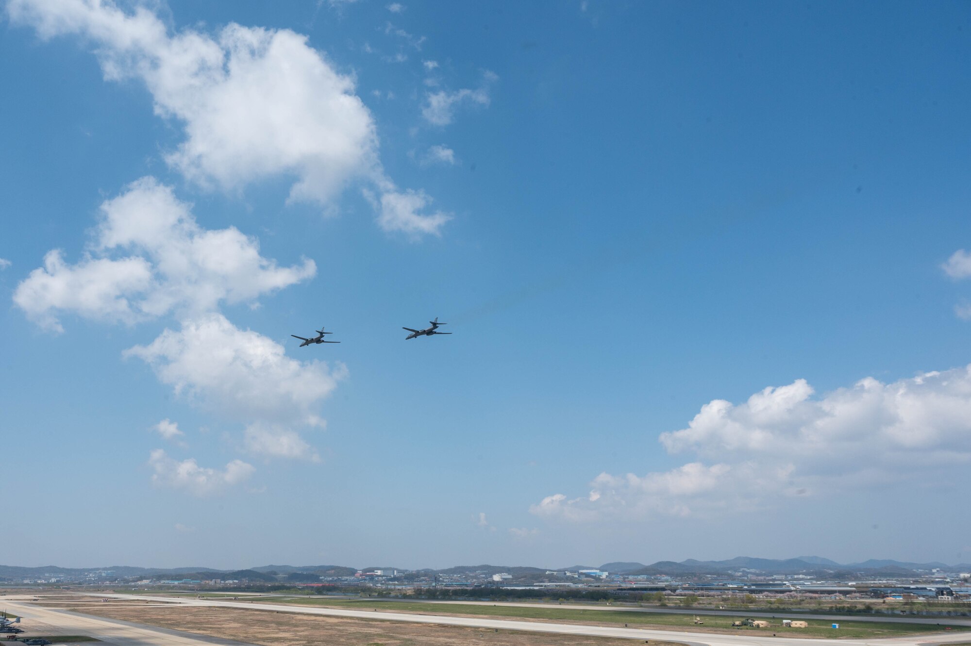 Two B-1 bomber aircraft fly close together in a blue sky with a runway blue and town in the distant background.