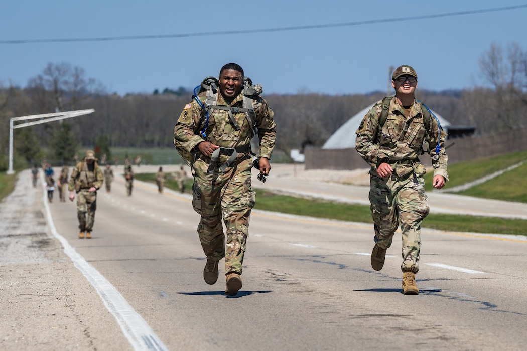 Two military members in uniform run towards the viewer with other participants visible in the distance