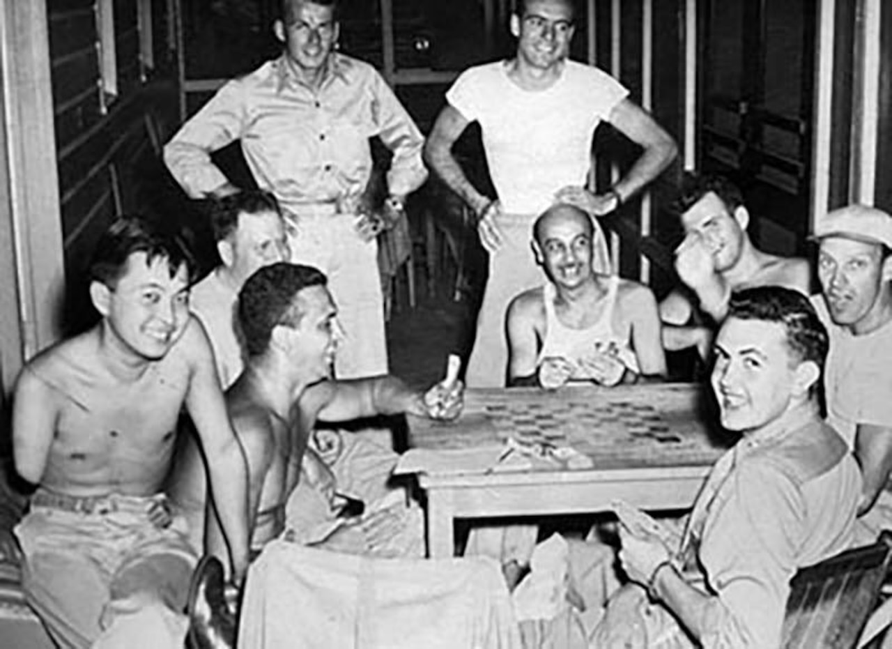 Nine men sitting and standing around a game table smile and laugh.