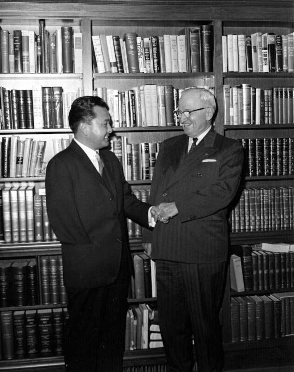 Two men shake hands in front of a wall of books.