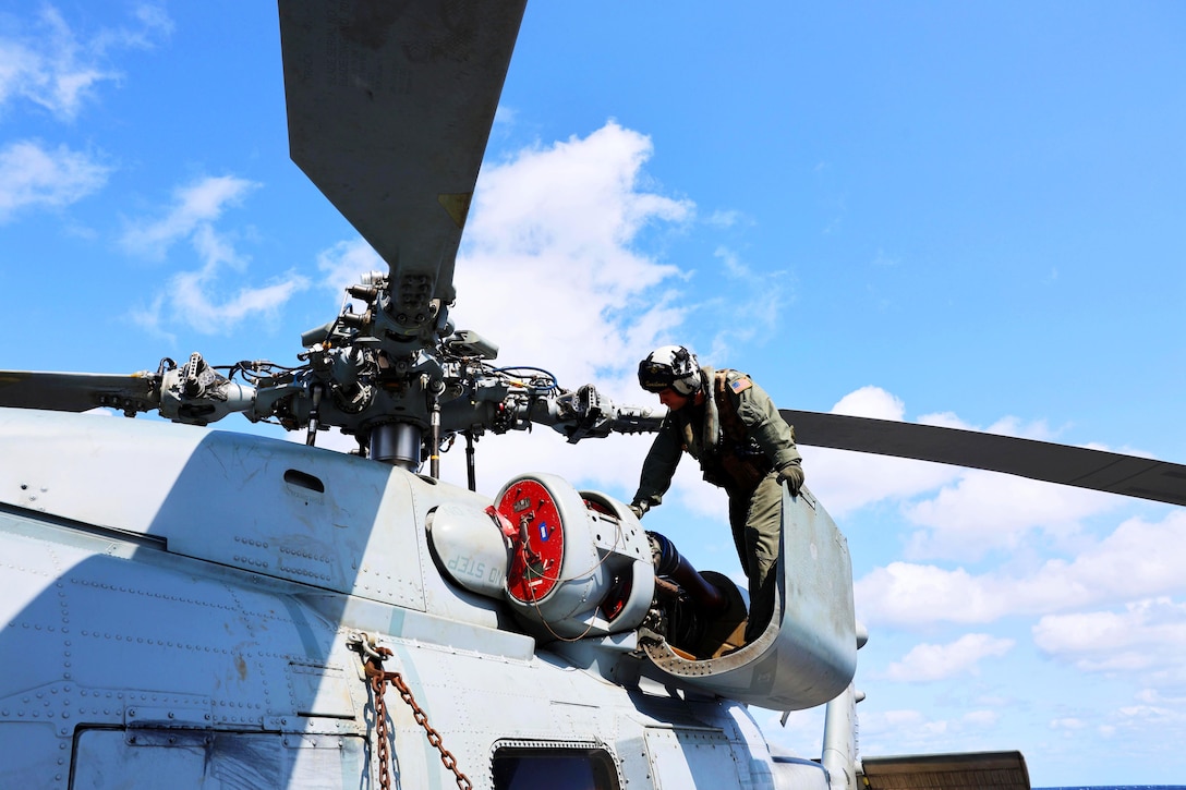 A sailor works on a helicopter against a cloudy, blue sky.