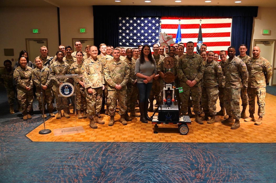 military members posed in front of a sculpture with many concoctions on it made by various squadrons, u.s. flag in background of people