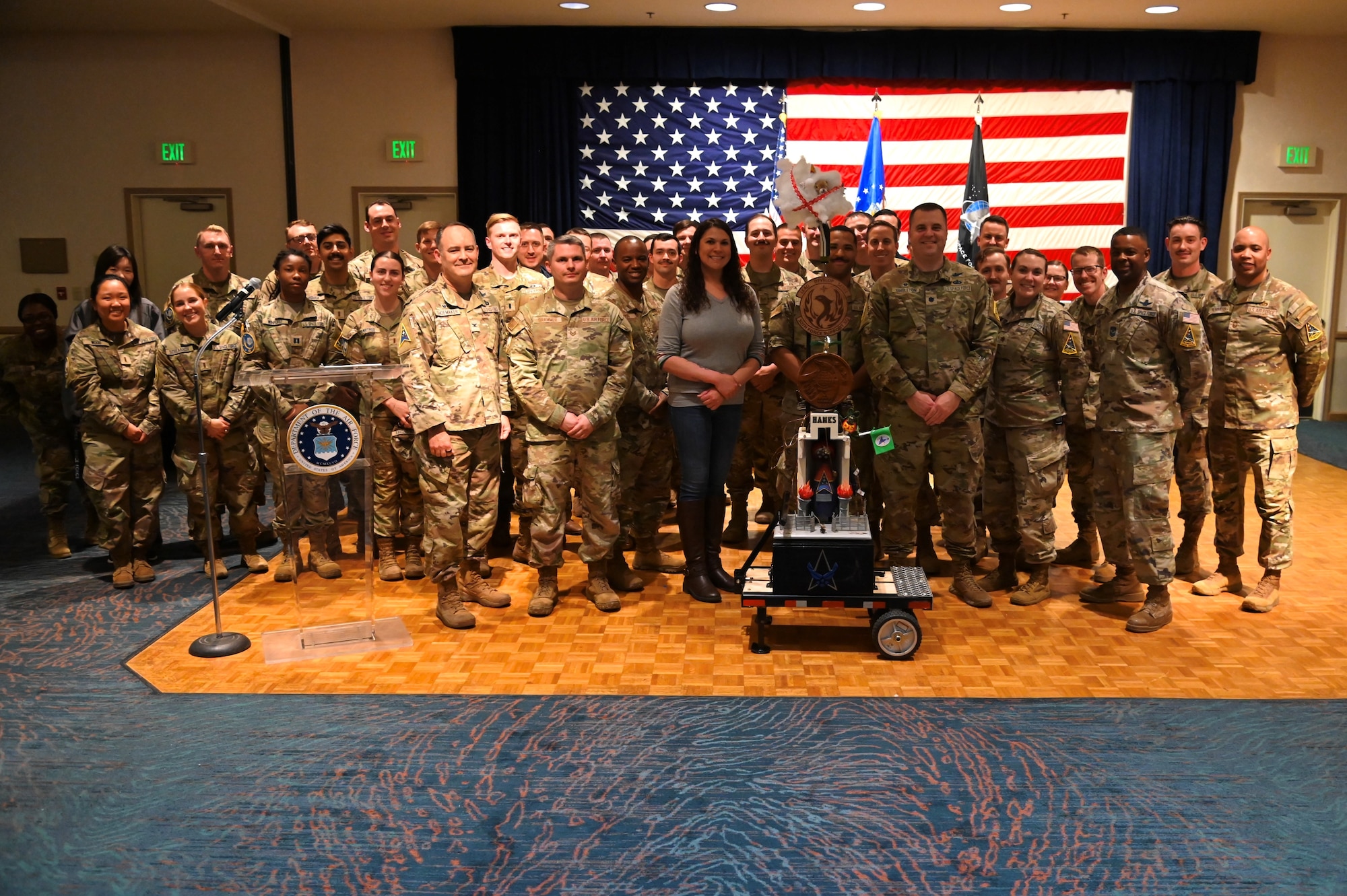 military members posed in front of a sculpture with many concoctions on it made by various squadrons, u.s. flag in background of people