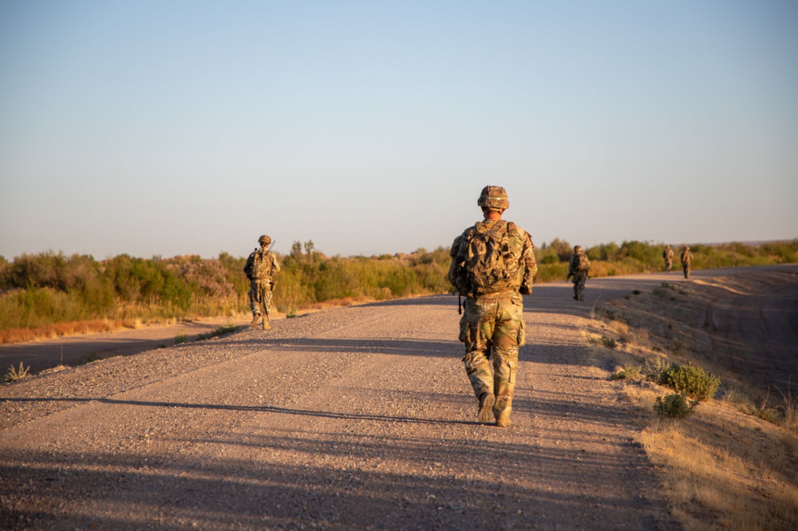 Four people in camouflage uniforms walk along a dusty road.