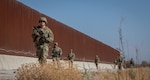 Eight people in camouflage uniforms walk along a gravel road with a large fence in the background.