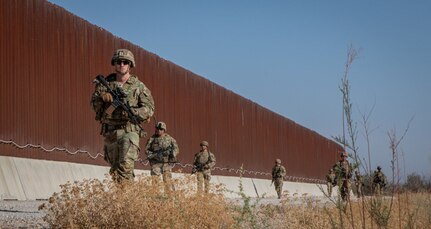 Eight people in camouflage uniforms walk along a gravel road with a large fence in the background.