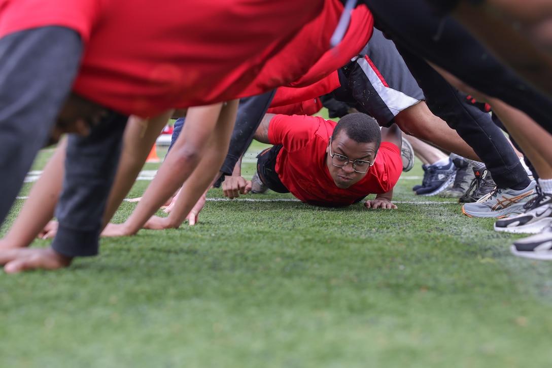 A Marine Corps poolee in a red T-shirt crawls under fellow poolees creating a human tunnel on a field.