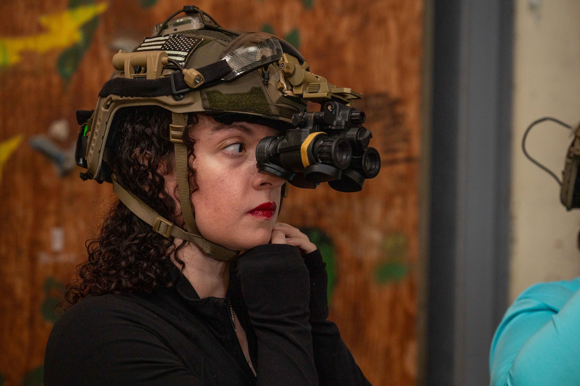 A woman wears a military helmet and looks through the night vision goggles mounted on the helmet.