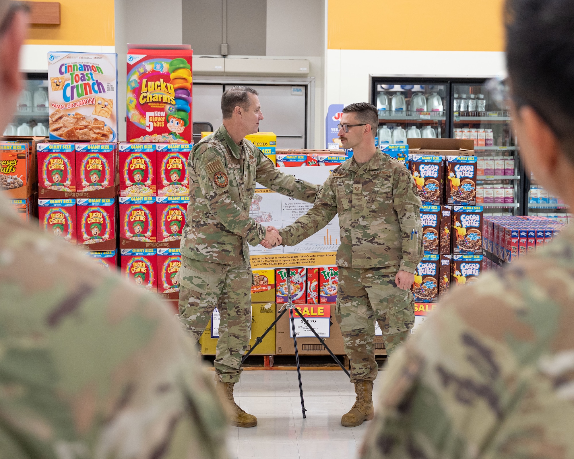 Two service members shake hands in front of food pallets