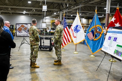 Two men wearing camouflage military uniforms stand in a warehouse talking.