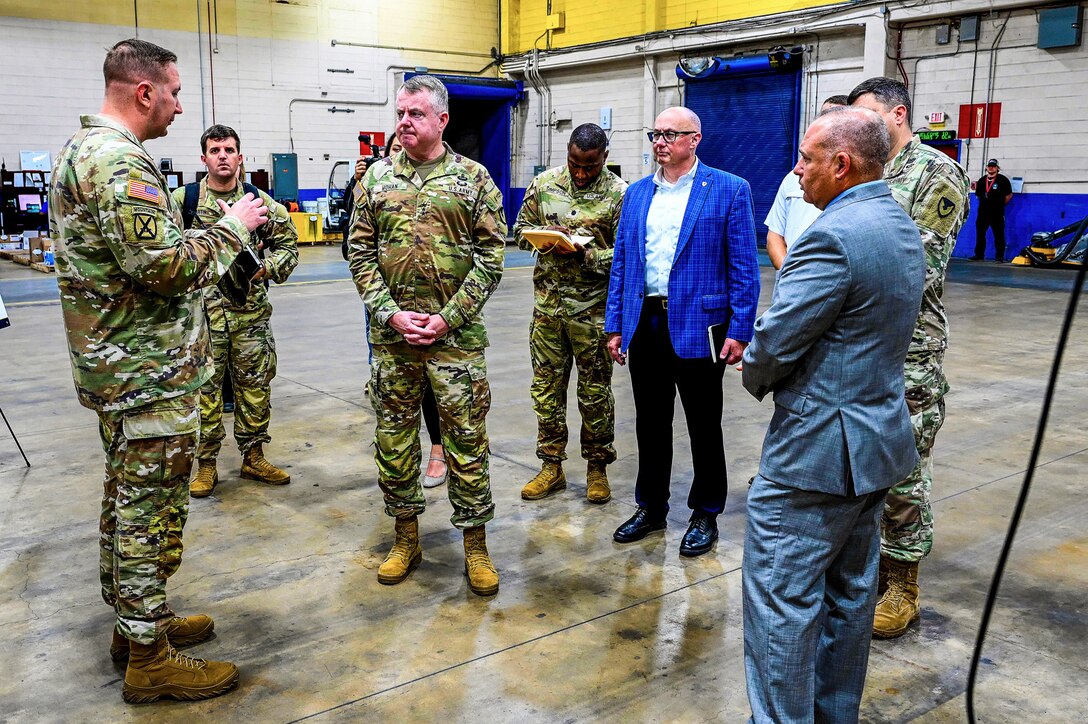 A group of men, several wearing camouflage military uniforms, stand in a warehouse talking.