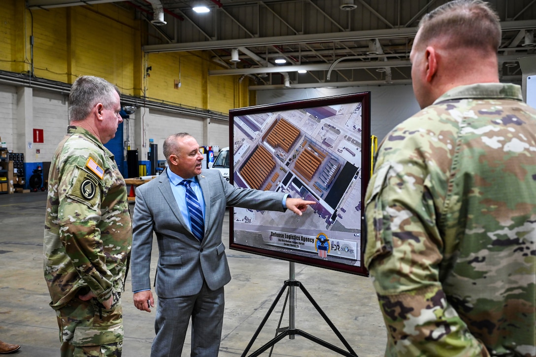 A man standing in a warehouse points to a display board as two men wearing camouflage military uniforms look on.