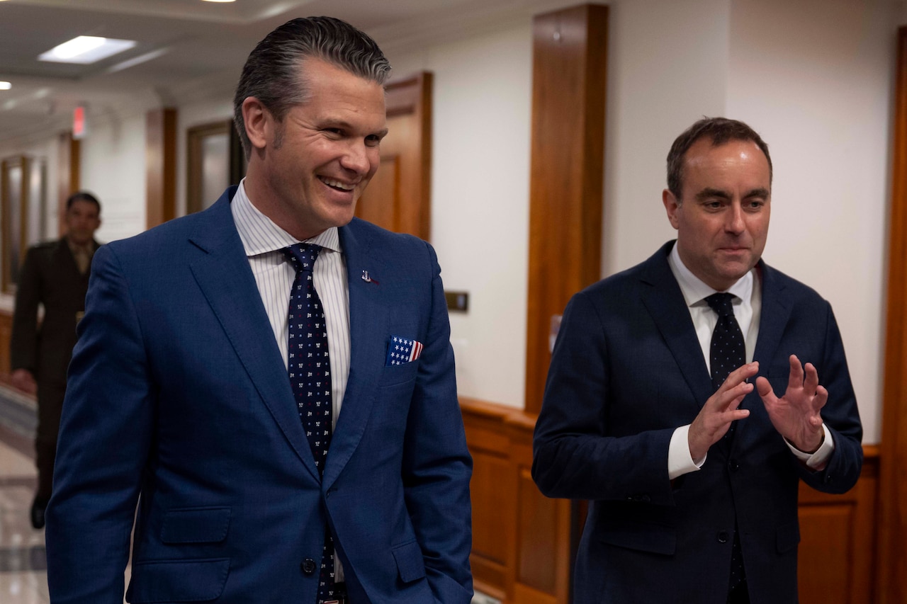Two men in suits walk down a hallway and talk.