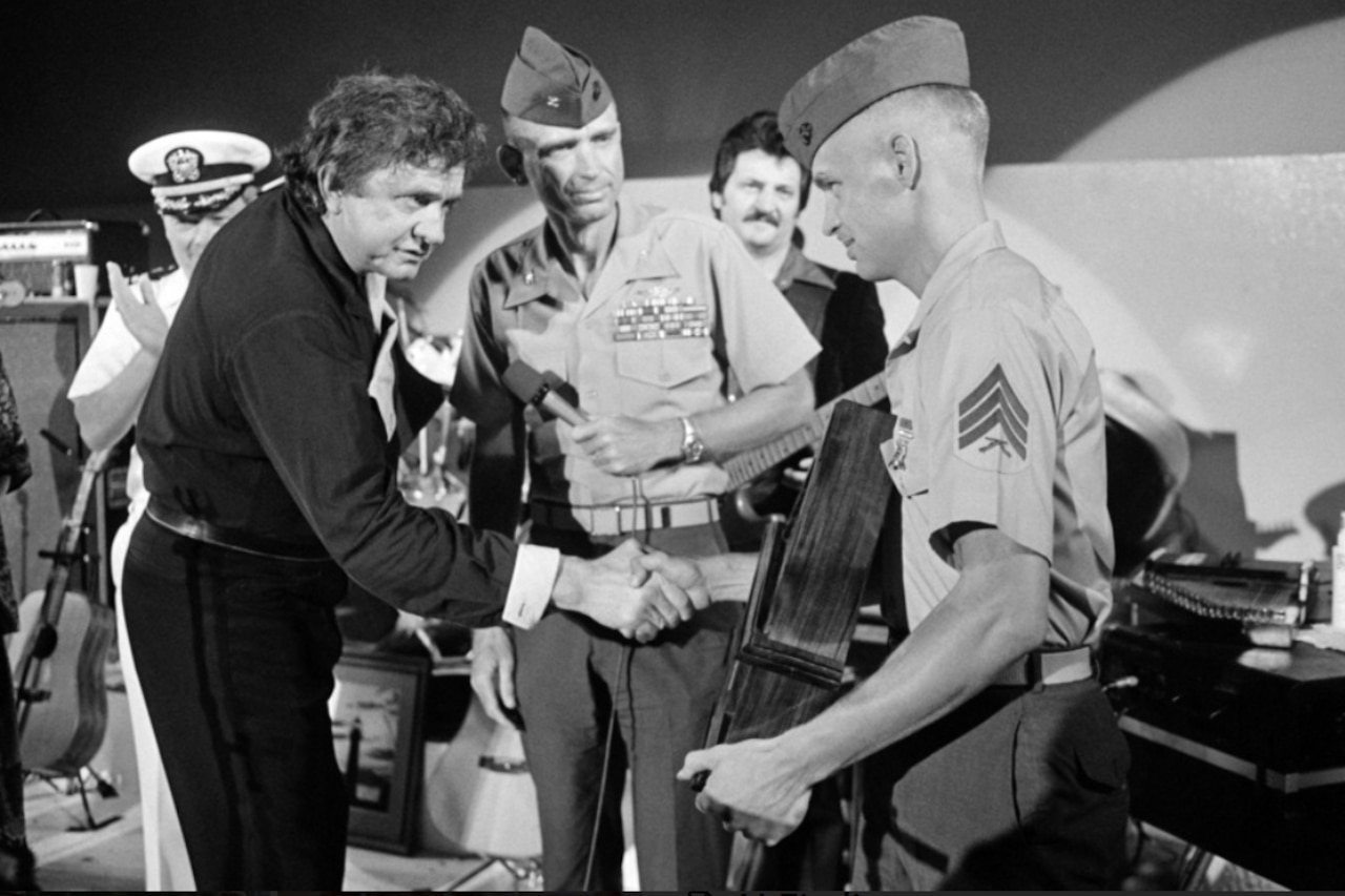 A man in business attire, left, shakes hands with a man in a military uniform. Another man in a military uniform stands behind them, holding a microphone.
