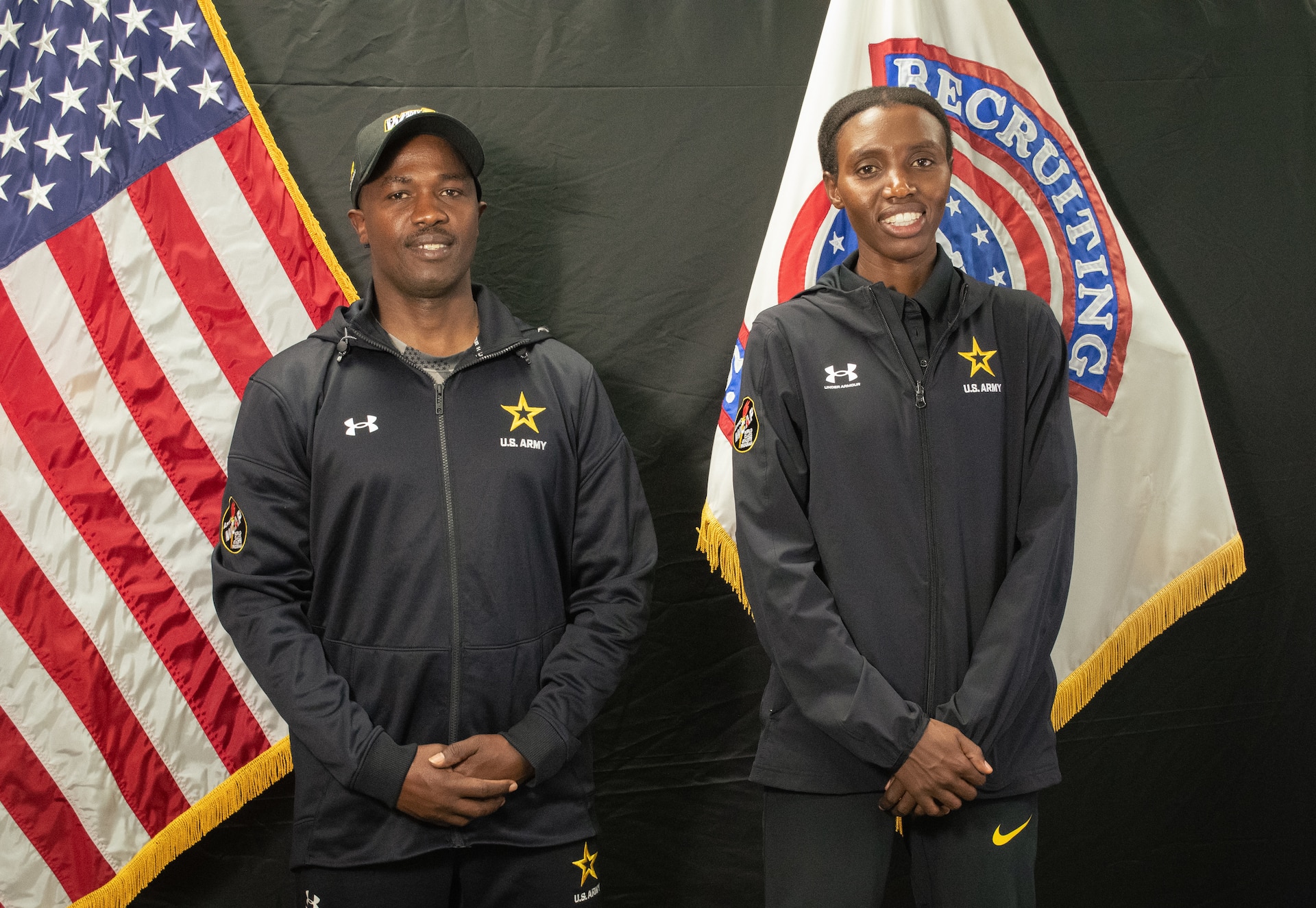 Male Soldier and female Soldier wearing Army branded track suits pose in front of U.S. National and Army Recruiting Command flags.