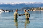 Two sailors shake hands while standing in the ocean with mountains in the distance.