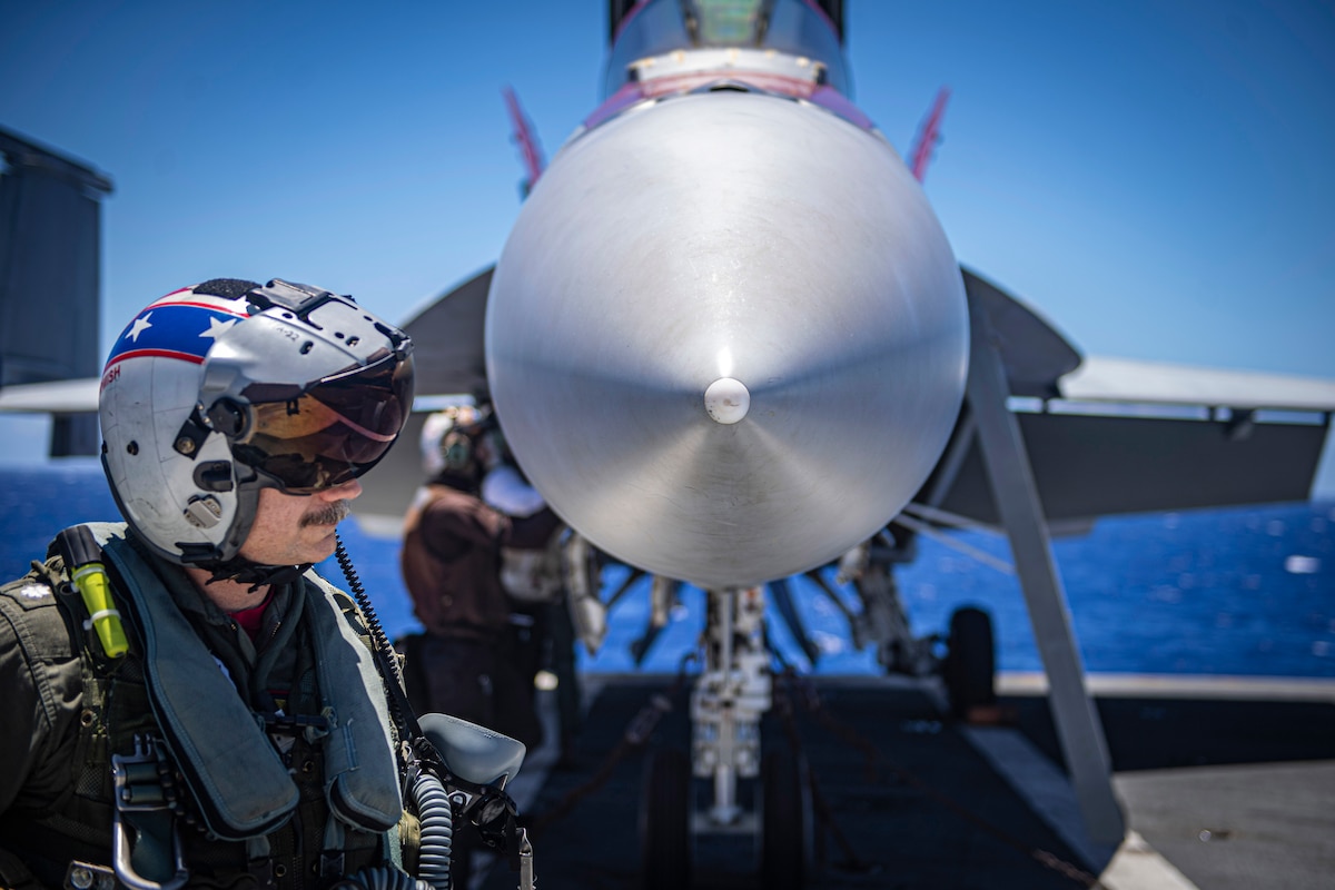 A sailor in a helmet stands next to the front of a silver aircraft.