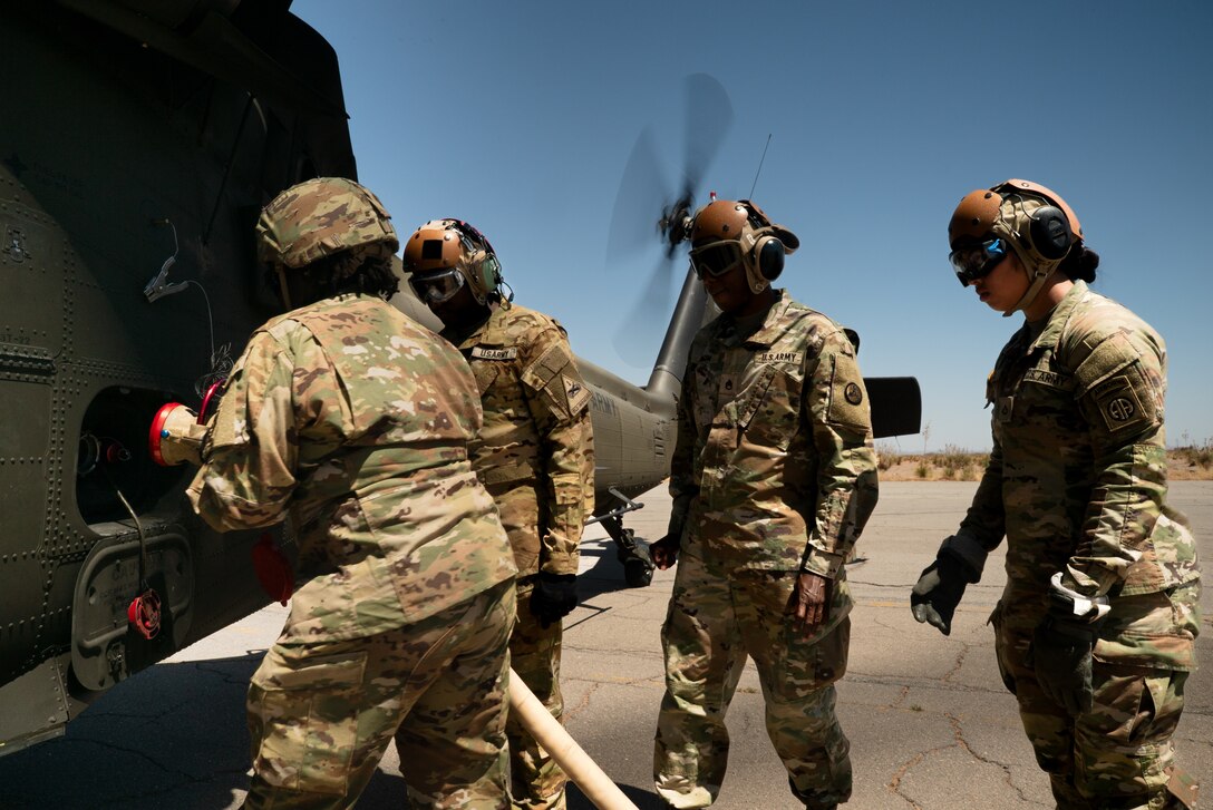 Two soldiers hook up hoses to refuel a helicopter while two soldiers observe.