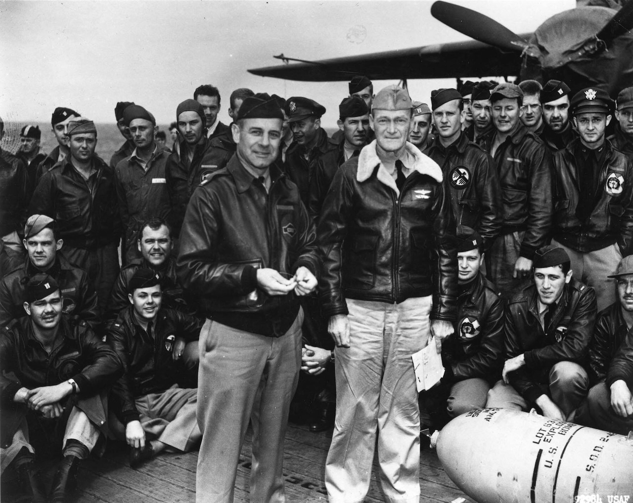 Two men in flight jackets stand on an aircraft carrier deck while large group of men, also wearing flight jackets stand and kneel behind them.