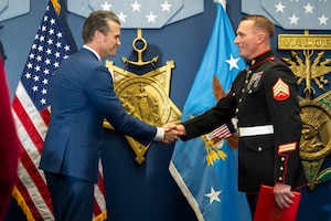 Two men shake hands in front of wall covered in medals.