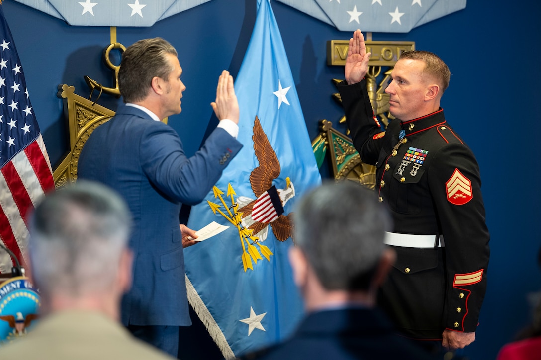 Two men raise their right hands while facing each other in front of a decorated wall.