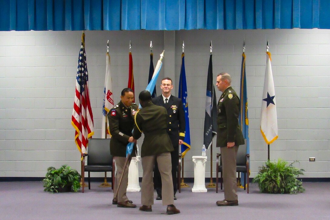 Four men stand on a stage in an indoor auditorium wearing military uniforms. One of them is passing a guidon to another.