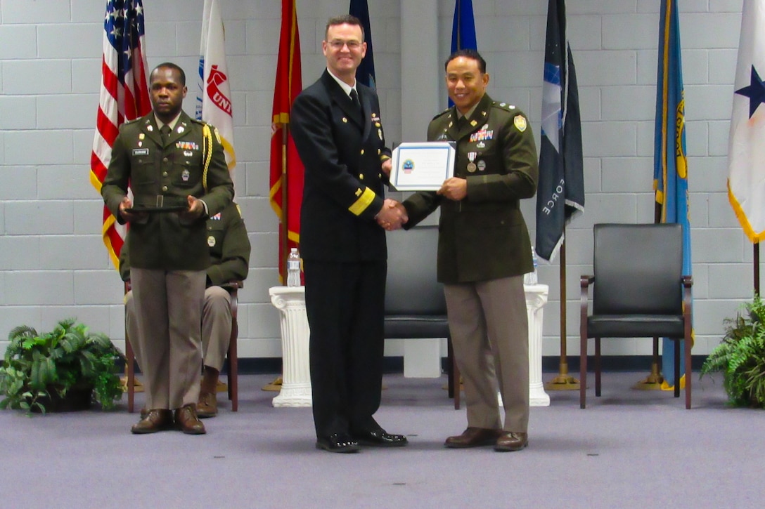 Three men stand on a stage in an indoor auditorium wearing military uniforms. Two of them are shaking hands and holding a certificate.