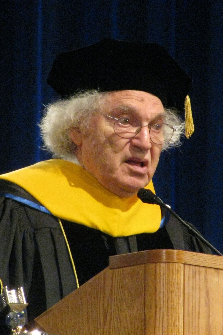 A man in a scholarly robe speaks at a lectern.