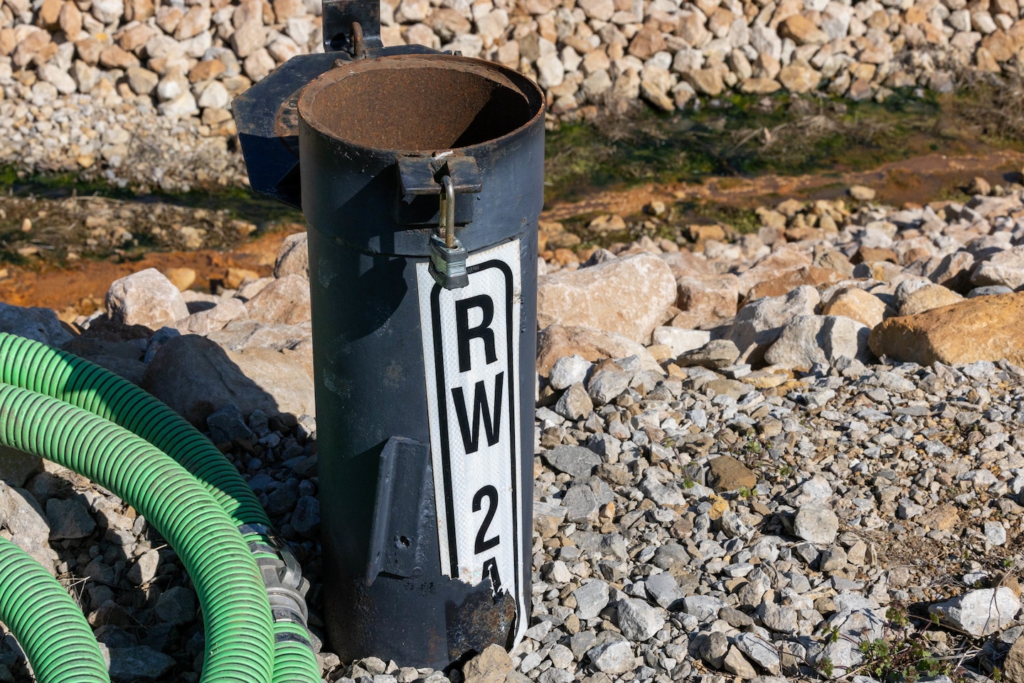 A metal tube sticks out of the ground with a green hose to the left and rocks behind.