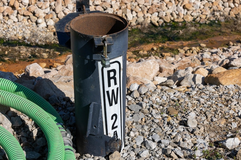 A metal tube sticks out of the ground with a green hose to the left and rocks behind.