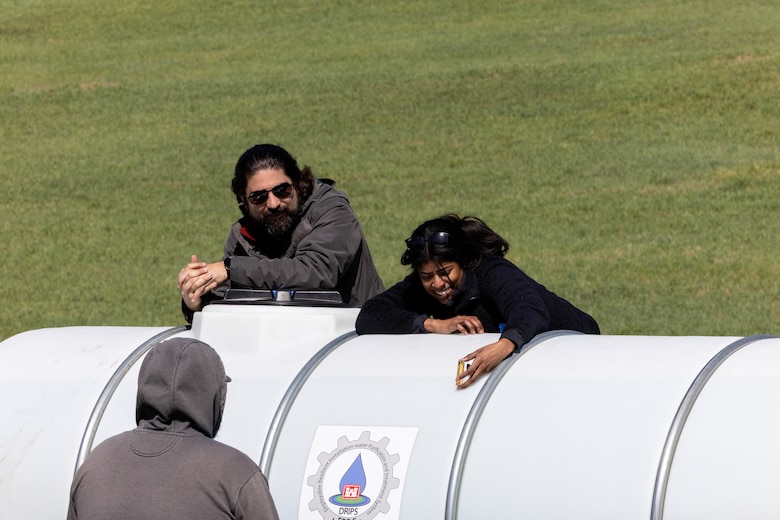 A man in a grey shirt and a woman in a black shirt show a man in a grey shirt a test tube while standing above a white plastic water tank.