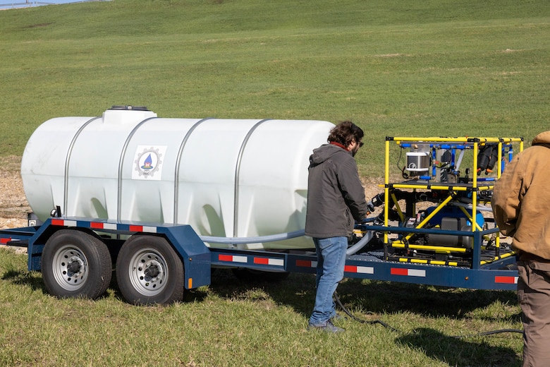 A large white water tank sits on a trailer and a man in a grey sweatshirt stands next to a large filtration system.