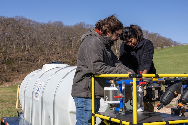 A man in a grey sweatshirt and a woman in a black shirt work on a large filtration system with trees in the background.