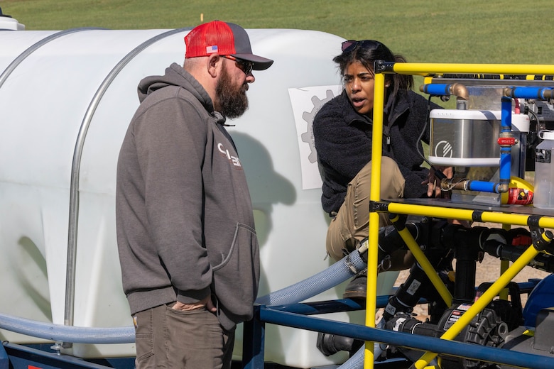 A man in a red hat and grey sweatshirt listens as a woman in a black shirt teaches him how to use a filtration machine.