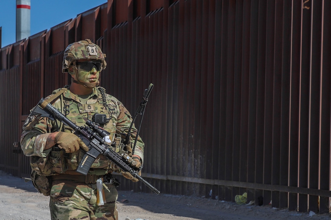 A soldier holding a weapon patrols next to a large wall.