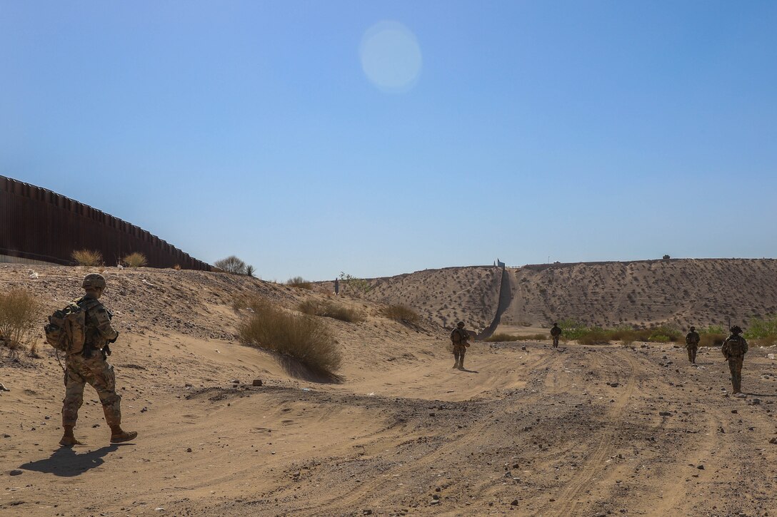 Soldiers patrol along desert terrain near a large wall.