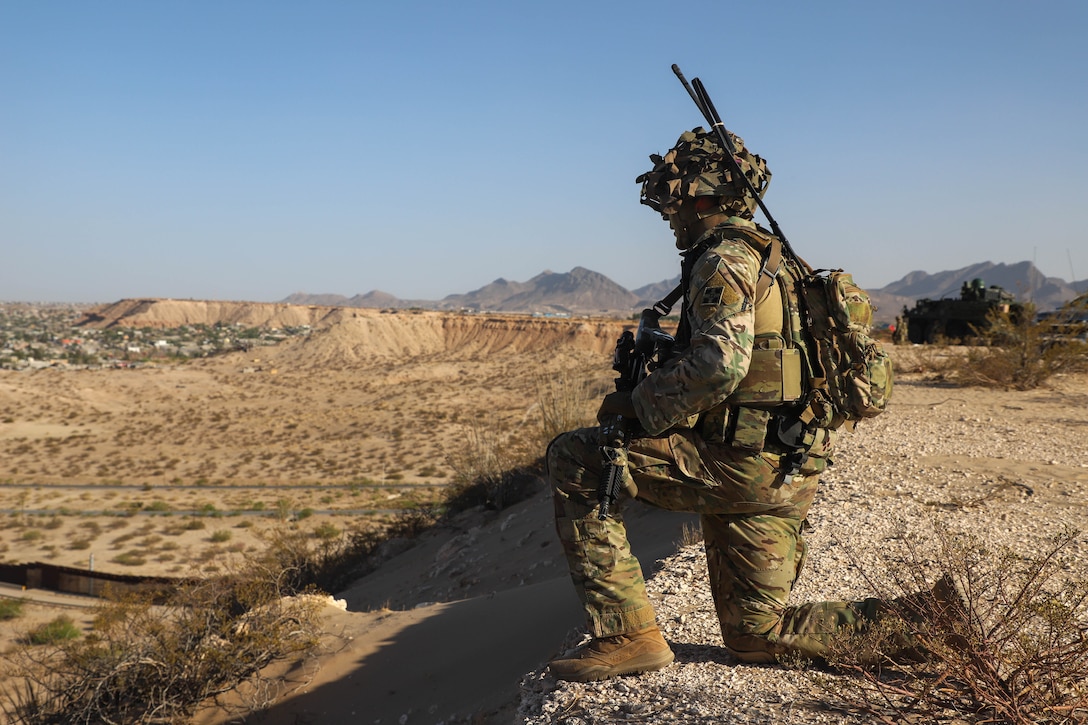 A soldier knees atop a hill over looking a desert valley during the day.
