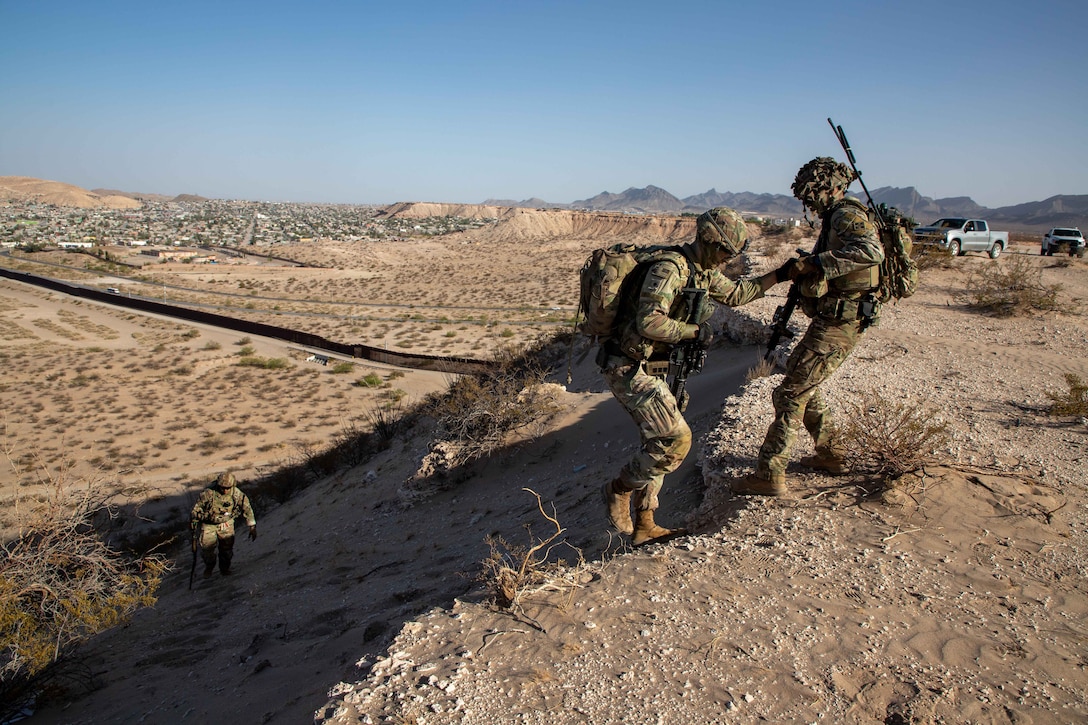 A soldier gives a hand to another soldier climbing up a hill during the day in the desert.