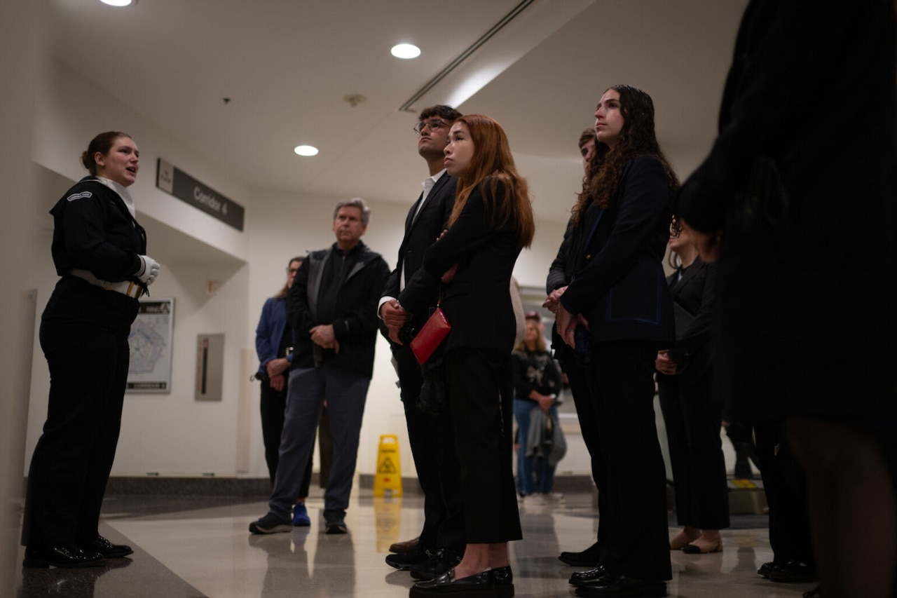 A woman in dress military uniform is standing while addressing a group of young people in civilian clothes who are also standing.