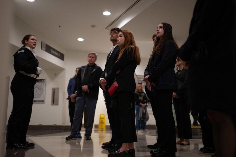 A woman in dress military uniform is standing while addressing a group of young people in civilian clothes who are also standing.