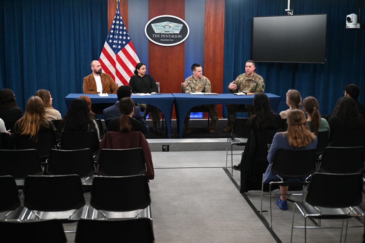 Four people, two of them in camouflage military uniforms and two in civilian attire, are sitting behind a table on a stage addressing a group of people seated in chairs. Behind the table is an American flag and a sign on the wall that reads “The Pentagon.”
