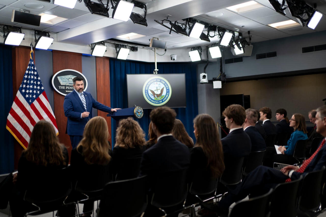 A man in a business suit is standing near a podium on stage talking to a group of young men and women sitting in chairs in front of him.