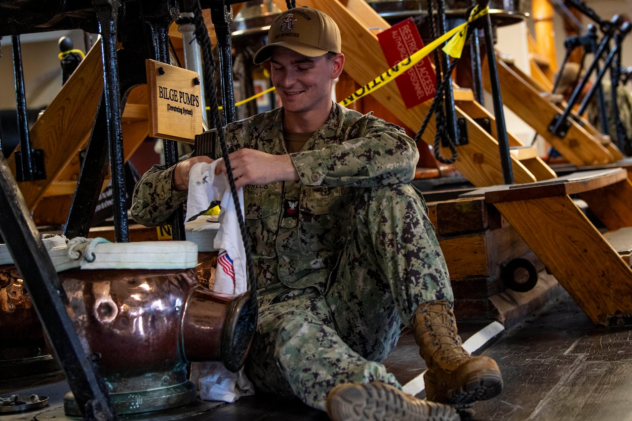 A sailor smiles and polishes machinery on an old ship while sitting on the ground.